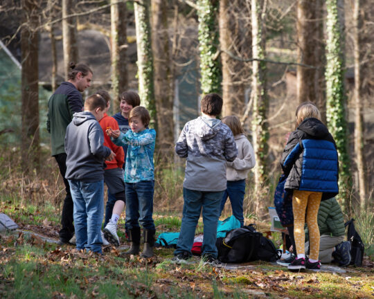 Students standing in front of a Muddy Sneakers Instructor a clearing in the woods, with backpacks and gear at their feet.