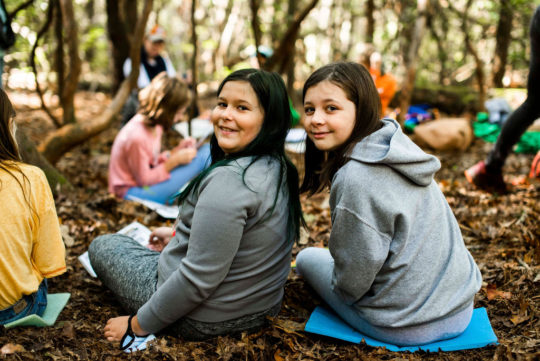 Two students sitting on leaf-covered ground with peers in the background, turn to smile at camera