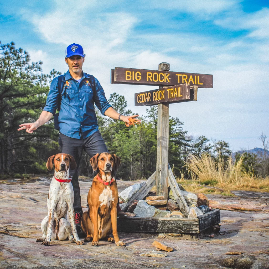 Robert Prioleau stands beside trail markers at the intersection of Big Rock Trail and Cedar Rock Trail, with two dogs sitting by his feet.