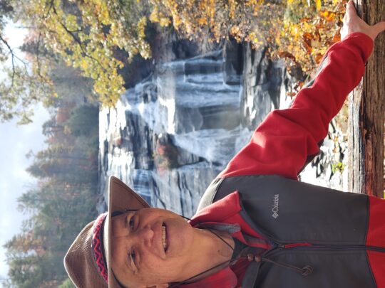 Muddy Sneakers Board Member Morris Jenkins stands on a wooden platform in front of a waterfall, beside colorful fall foliage