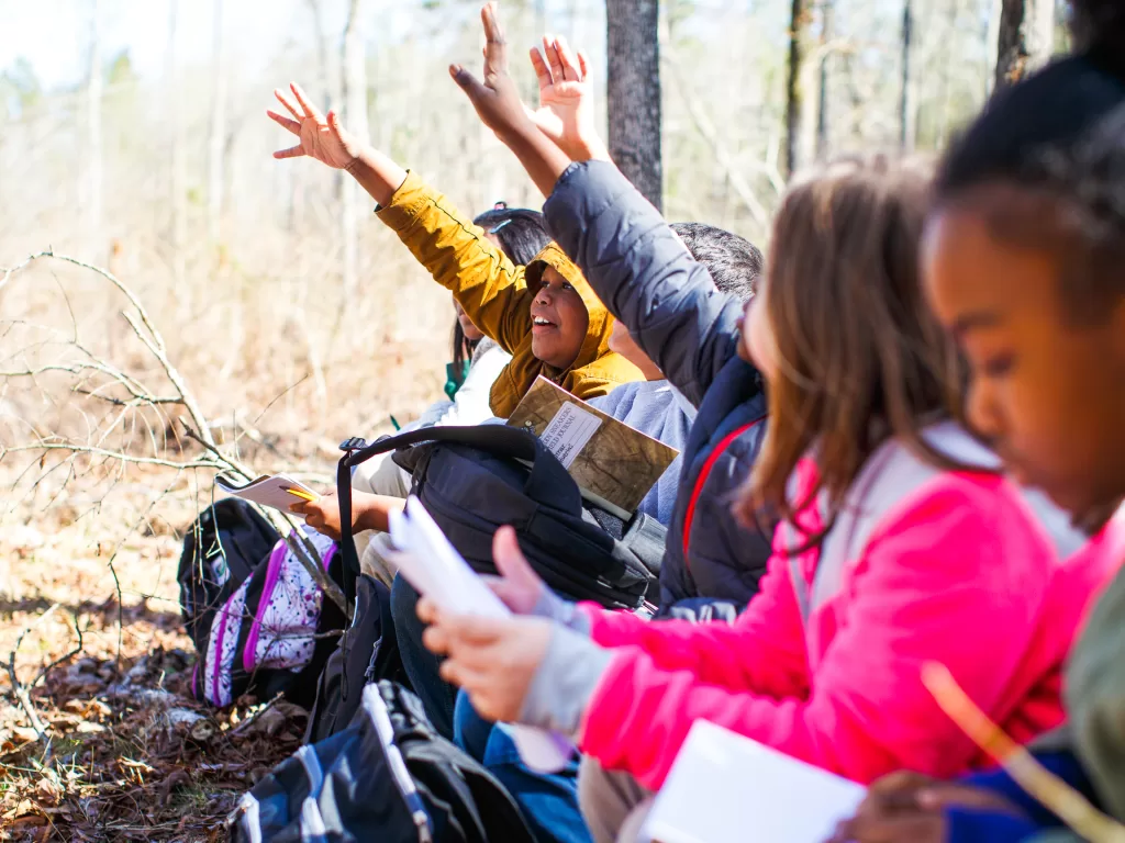 Students with big smiles on their faces, raise their hands enthusiastically as they participate in a Muddy Sneakers lesson in the woods.