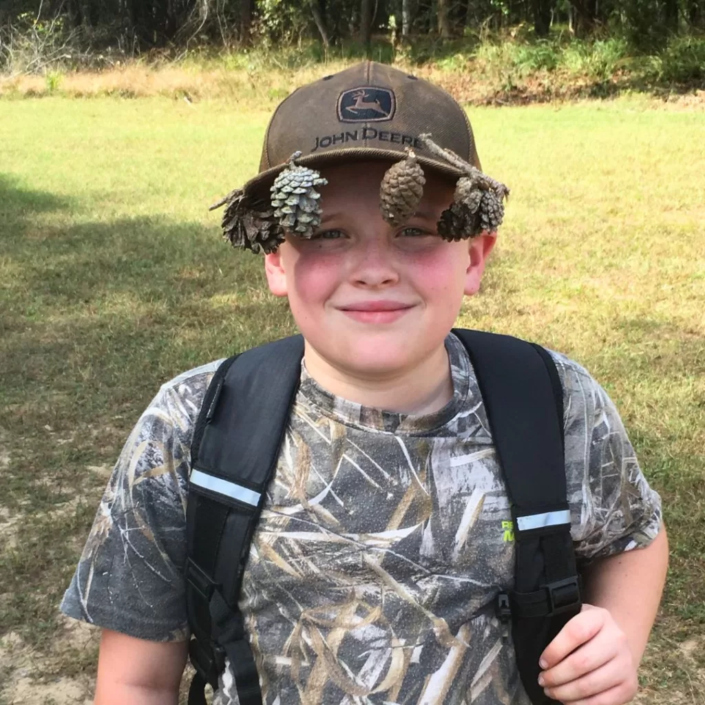 Student smiles at the camera, showing off his creativity, one of the many benefits of outdoor education, by hanging pinecones from the rim of his hat.