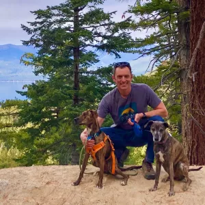 Glenn Middleton, Muddy Sneakers' Director of Programming, poses for a photo alongside his two dogs along a rocky trail, with a lake and mountains in the background