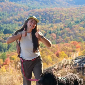 Emma Potter, Mountains Field Instructor, smiling at the camera from the trail, with a fall forested mountain in the background