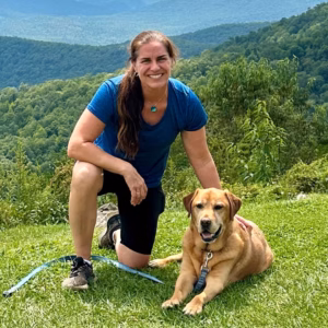 Gwynne Stump and her dog pose for a photo on a grassy mountaintop.