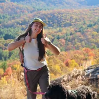 Emma Potter, Mountains Field Instructor, smiling at the camera from the trail, with a fall forested mountain in the background